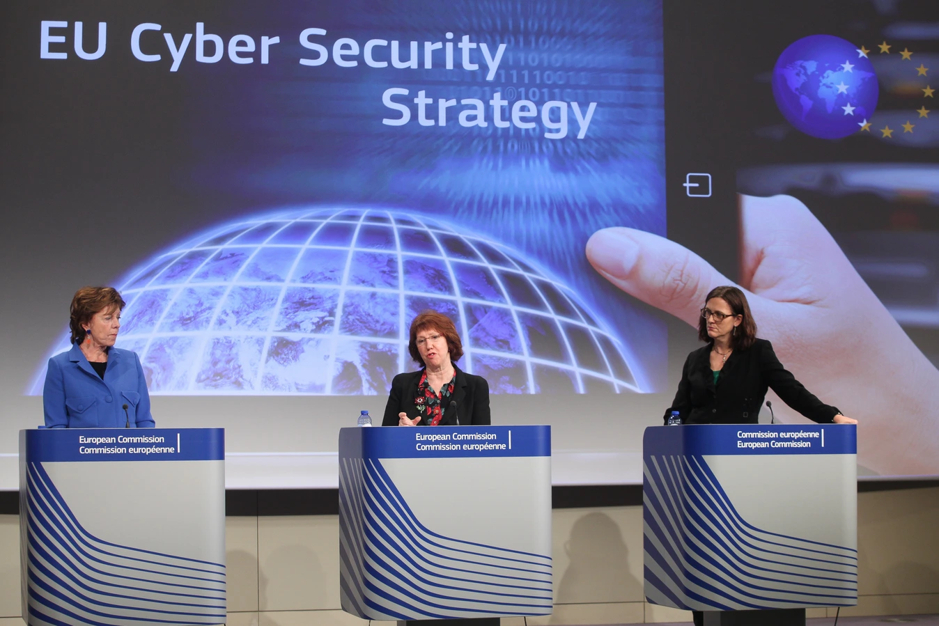 Press conference on the EU Cybersecurity Strategy, with three speakers at podiums in front of a screen showing the strategy.